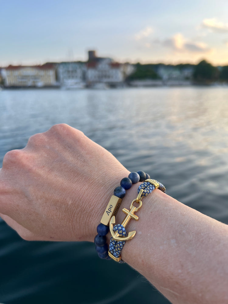Blue stone bead bracelet and gold anchor bracelet worn on a wrist above the water at Marstrand at sunset